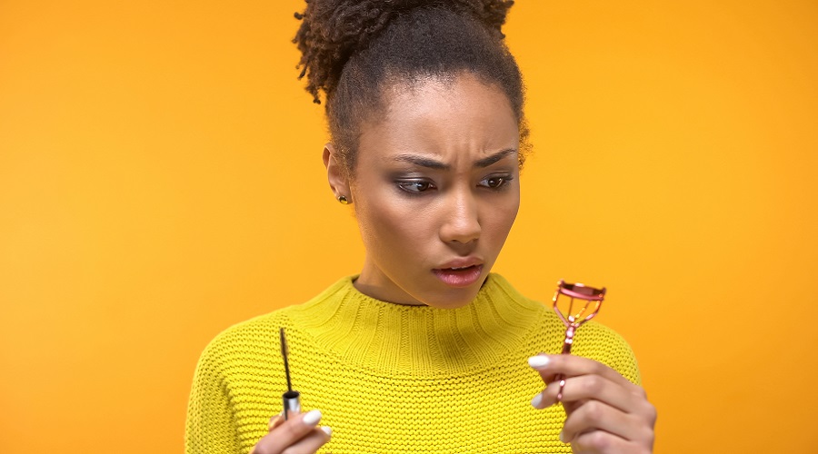 Confused black woman looking at mascara and eyelash curler, make-up tips, beauty