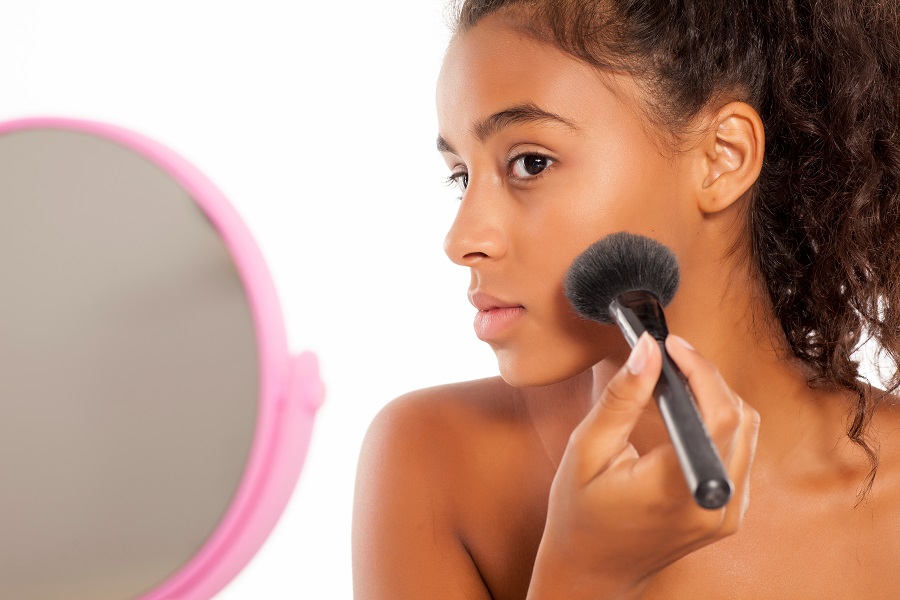 a young dark skinned woman applies a powder foundation on her fa
